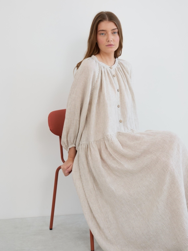 Woman seated in the Etta striped linen maxi dress with gathered round neckline, button-down front, and 3/4 balloon sleeves in beige and white