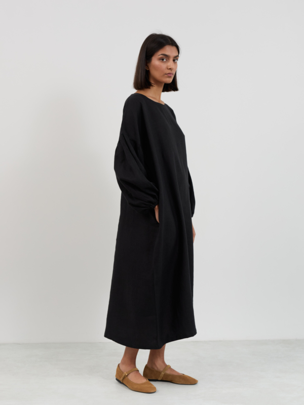 Three-quarter side view of the Briar linen midi dress in black, hand in pocket, showing balloon sleeves and relaxed straight silhouette