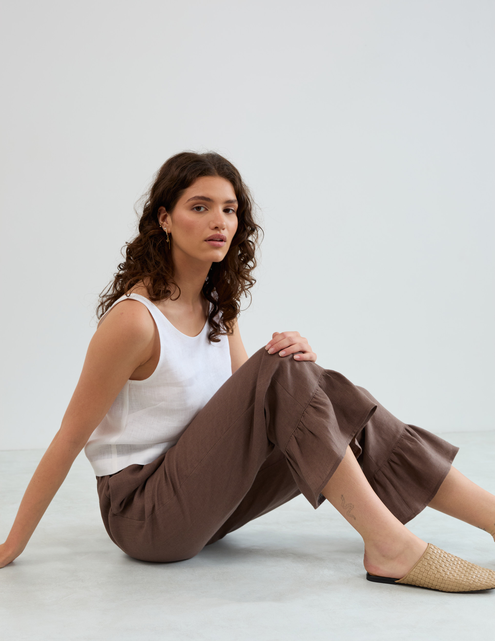 Model seated on floor wearing Gigi cocoa linen ruffle hem pants with a white linen tank top and woven mules